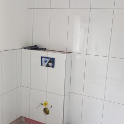 Kudzia Pawel - Białe, błyszczące płytki w łazience w trakcie wykańczania. Widoczne przyłącza do toalety podtynkowej i fragment podłogi przed położeniem płytek. Minimalistyczny design.