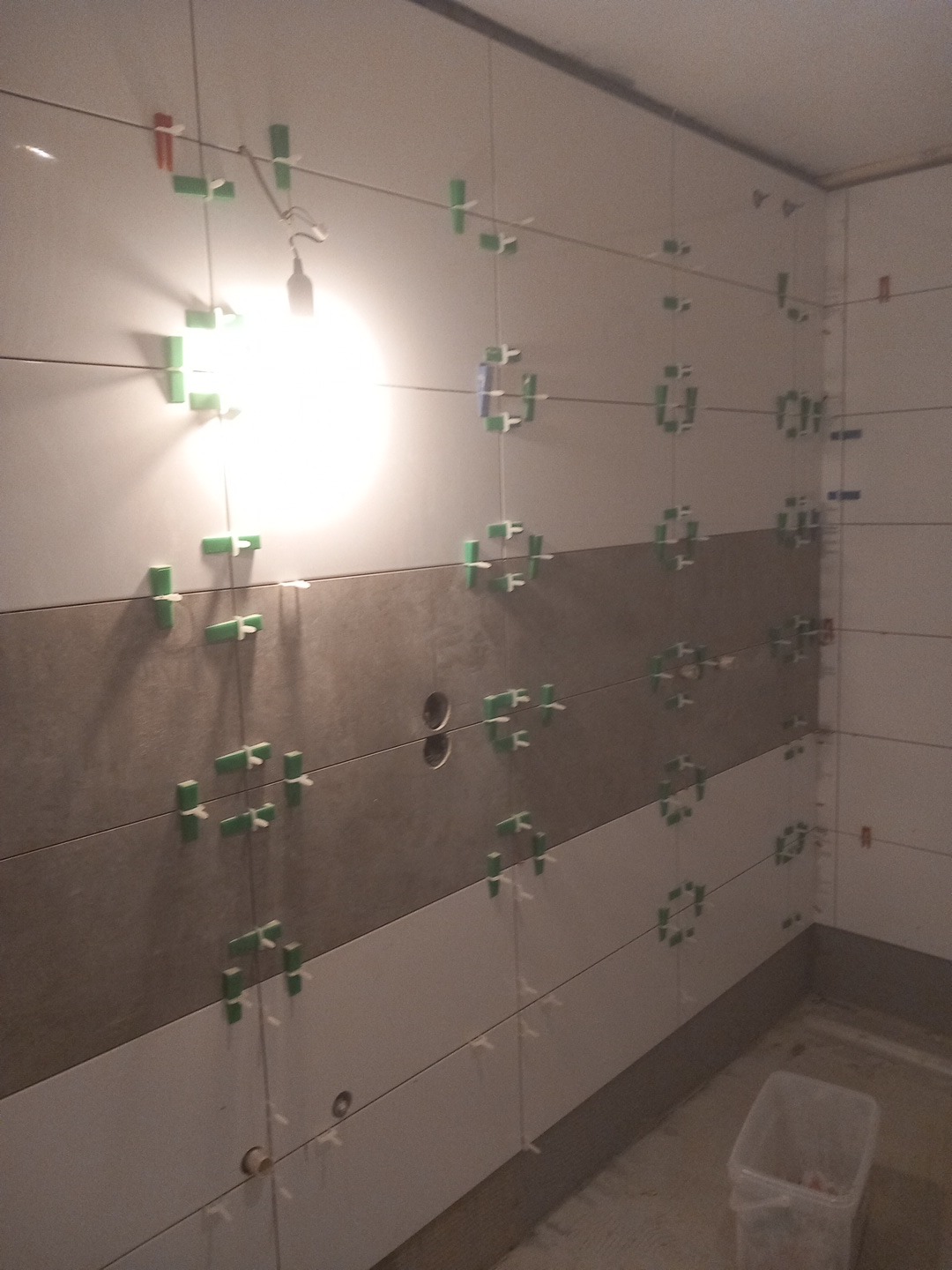 Ściana łazienki w trakcie układania płytek, widoczne krzyżyki dystansowe i sznurki poziomujące. Część płytek w kolorze szarym, część białym. W tle widoczny fragment podłogi.