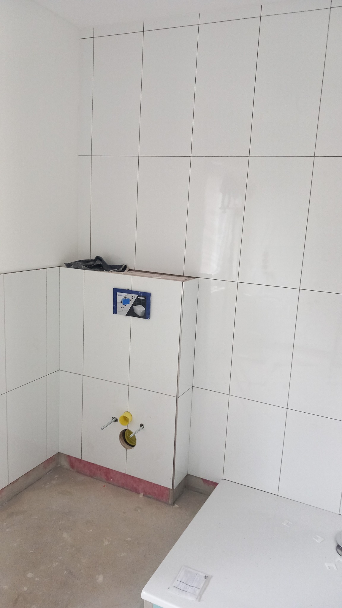 Białe, błyszczące płytki w łazience w trakcie wykańczania. Widoczne przyłącza do toalety podtynkowej i fragment podłogi przed położeniem płytek. Minimalistyczny design.