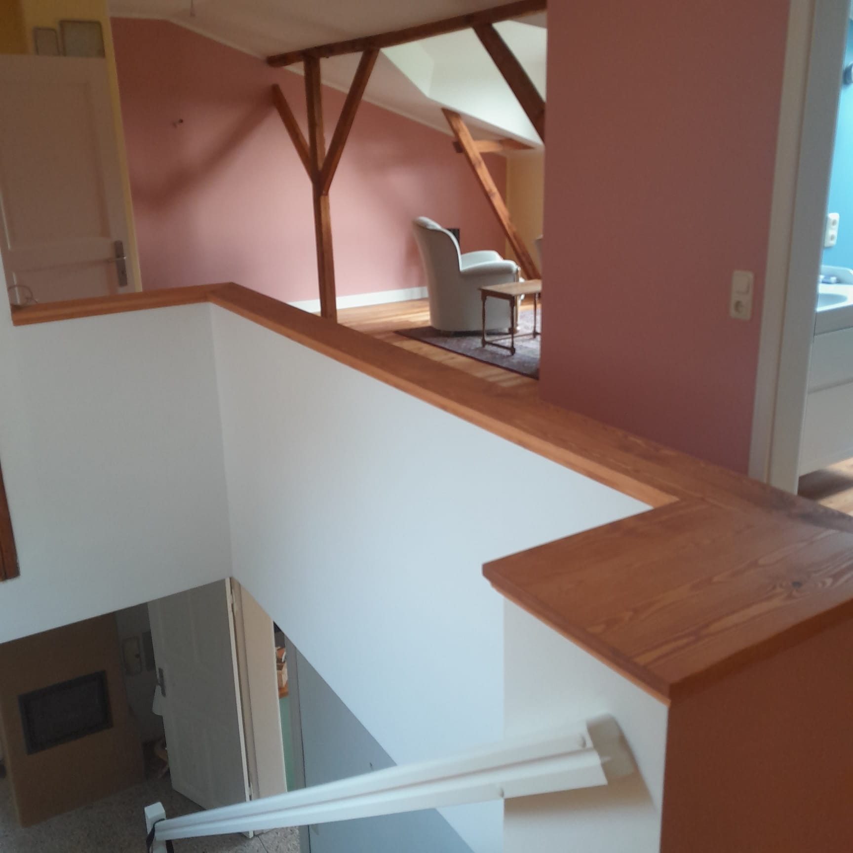 Jasne wnętrze domu z drewnianą konstrukcją stropu i białymi ścianami. Widoczny fragment schodów i pokój z fotelem. Ściany w odcieniach różu i błękitu.