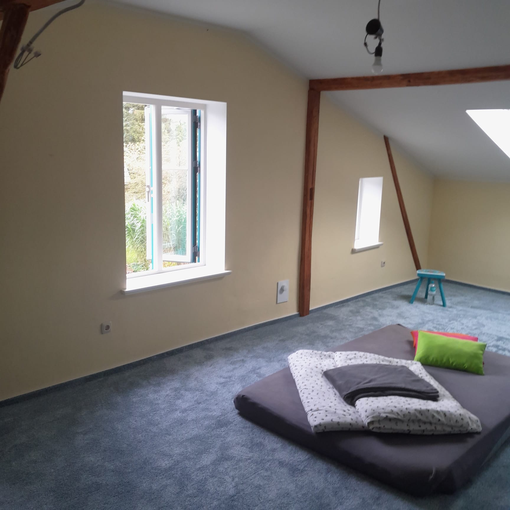 Jasne poddasze po remoncie: świeżo pomalowane ściany w odcieniach beżu, nowe okna z białymi ramami, szary dywan i drewniane elementy konstrukcyjne.