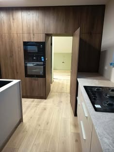 Nowoczesna kuchnia z zabudową w odcieniach brązu i beżu, z wbudowanym piekarnikiem i mikrofalówką. Widoczny fragment blatu i płyty grzewczej. Otwarty prześwit do innego pomieszczenia.