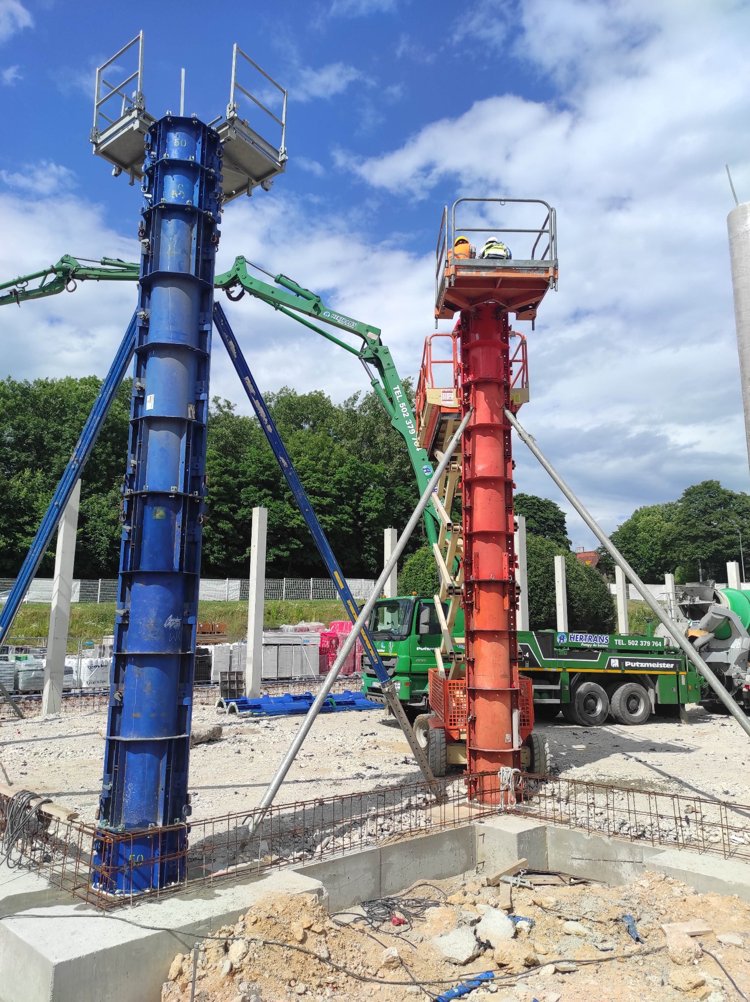 Kolorowe szalunki kolumnowe (czerwony i niebieski) z podestami roboczymi na budowie, wsparte na podporach. W tle betoniarka i pompy do betonu.