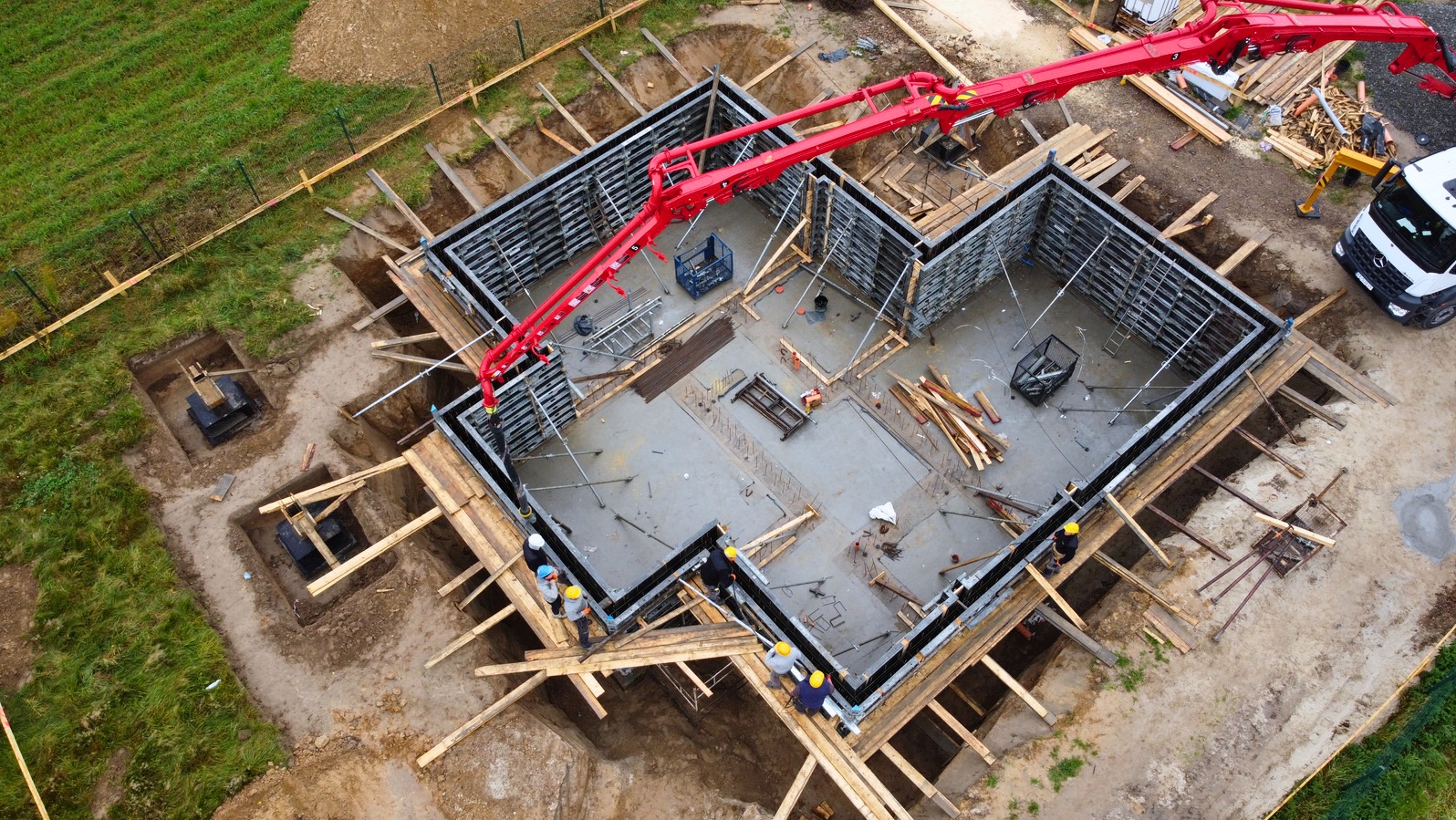 Z góry: wylewanie fundamentów domu z użyciem pompy do betonu. Widoczne szalunki, zbrojenia i pracownicy w kaskach. Częściowo widoczny samochód dostawczy.