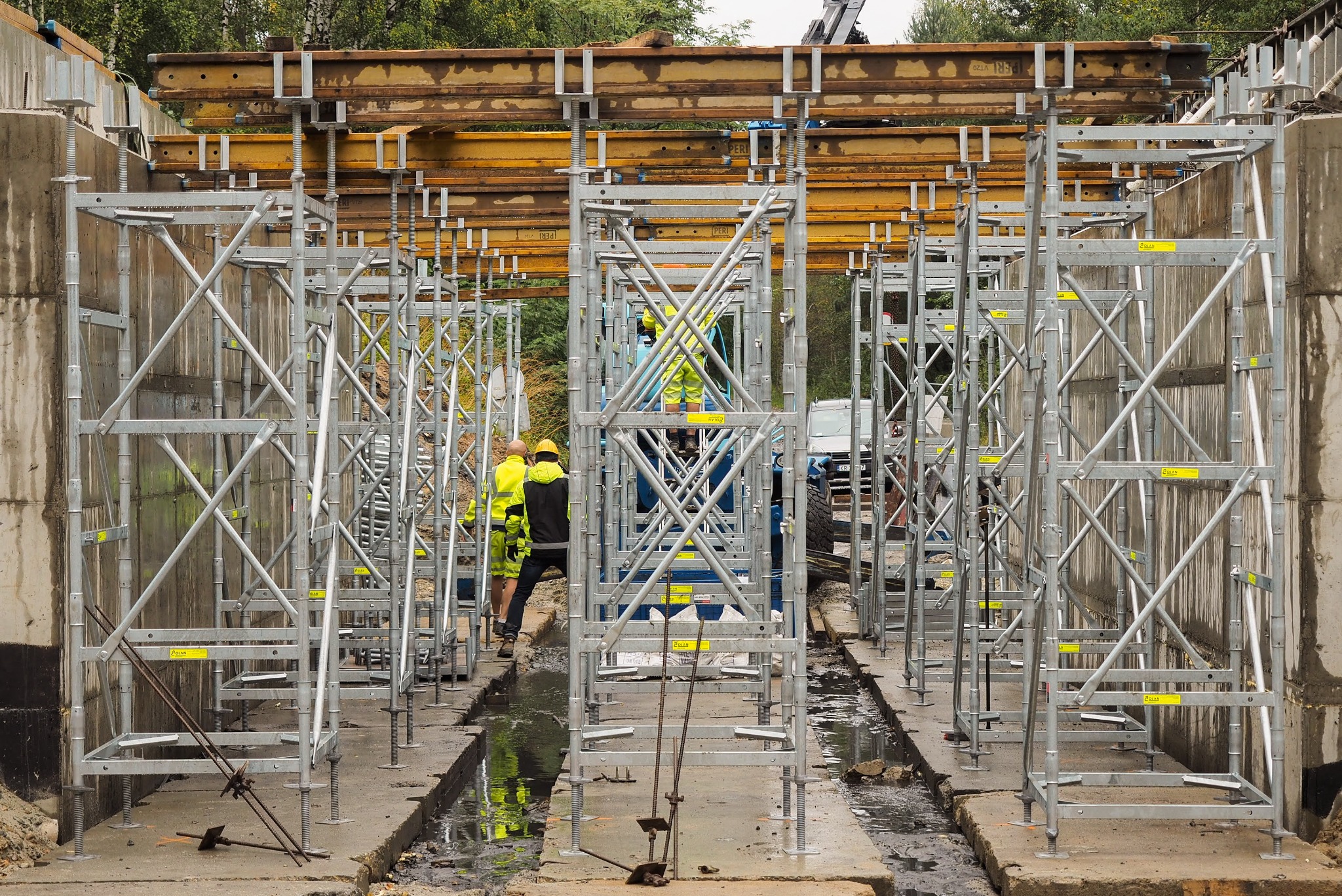 Szare, metalowe rusztowania budowlane z żółtymi elementami, ustawione w rzędach na placu budowy. W tle pracownicy w odblaskowych ubraniach i zaparkowany samochód.