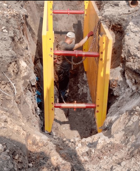 Montaż szalunku wykopu z żółtymi panelami i czerwonymi rozpórkami. Pracownik w kasku i rękawicach ustawia konstrukcję w wąskim wykopie.