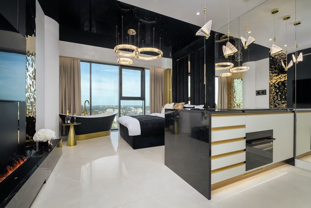 Luksusowe studio apartament z wanną przy oknie, łóżkiem i aneksem kuchennym. Czarno-złota kolorystyka, nowoczesne oświetlenie, widok na miasto.