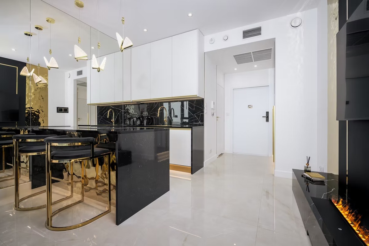 Nowoczesne wnętrze: czarna wyspa kuchenna z marmurowym blatem i złotymi detalami, białe szafki, lustrzana ściana, designerskie lampy i błyszcząca podłoga.
