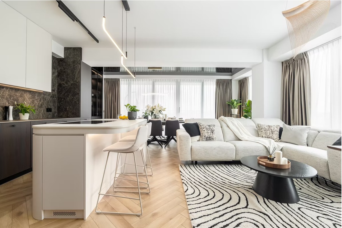 Jasne, przestronne wnętrze: kuchnia z wyspą, salon z sofą narożną i dywanem w paski. Nowoczesne oświetlenie, drewniana podłoga w jodełkę, duże okna z zasłonami.
