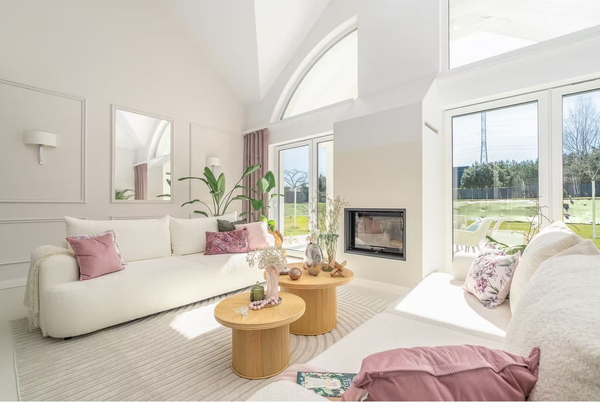 Jasne wnętrze salonu z kominkiem i dużymi oknami, miękkie sofy z poduszkami w odcieniach różu, okrągłe stoliki kawowe. Widok na zieleń za oknem.