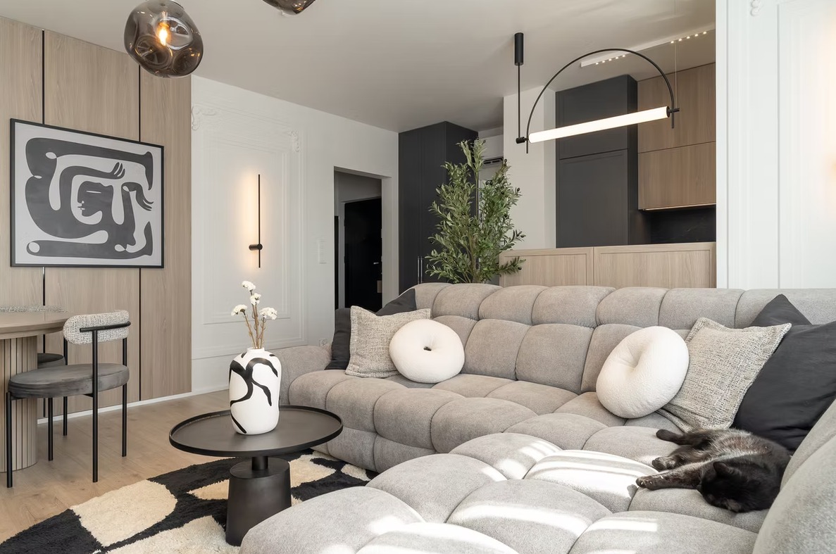 Stylowe wnętrze salonu z szarą sofą modułową, czarnym kotem, abstrakcyjnym obrazem i nowoczesnym oświetleniem. Widok na aneks kuchenny w tle.