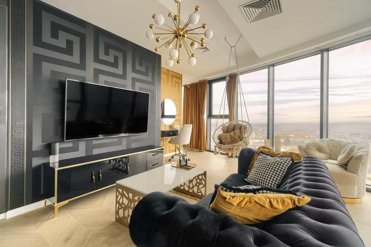 Elegancki salon z czarną sofą, wzorzystą tapetą i widokiem na miasto z wysokiego piętra. Lampa w stylu retro i wiszące krzesło dodają wnętrzu charakteru.