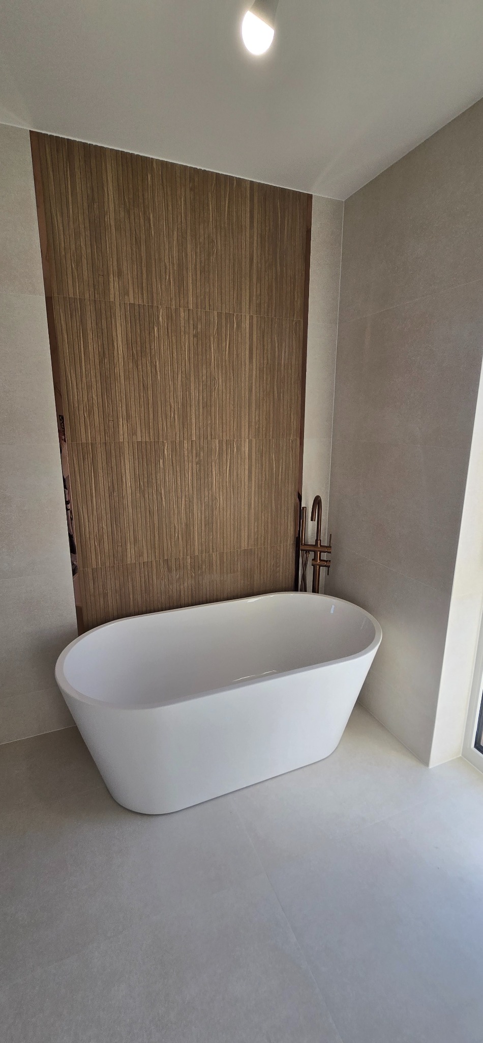 Nowoczesna łazienka z wolnostojącą białą wanną na tle ściany z drewnopodobnych paneli i szarej płytki. Minimalistyczny design, elegancka armatura.