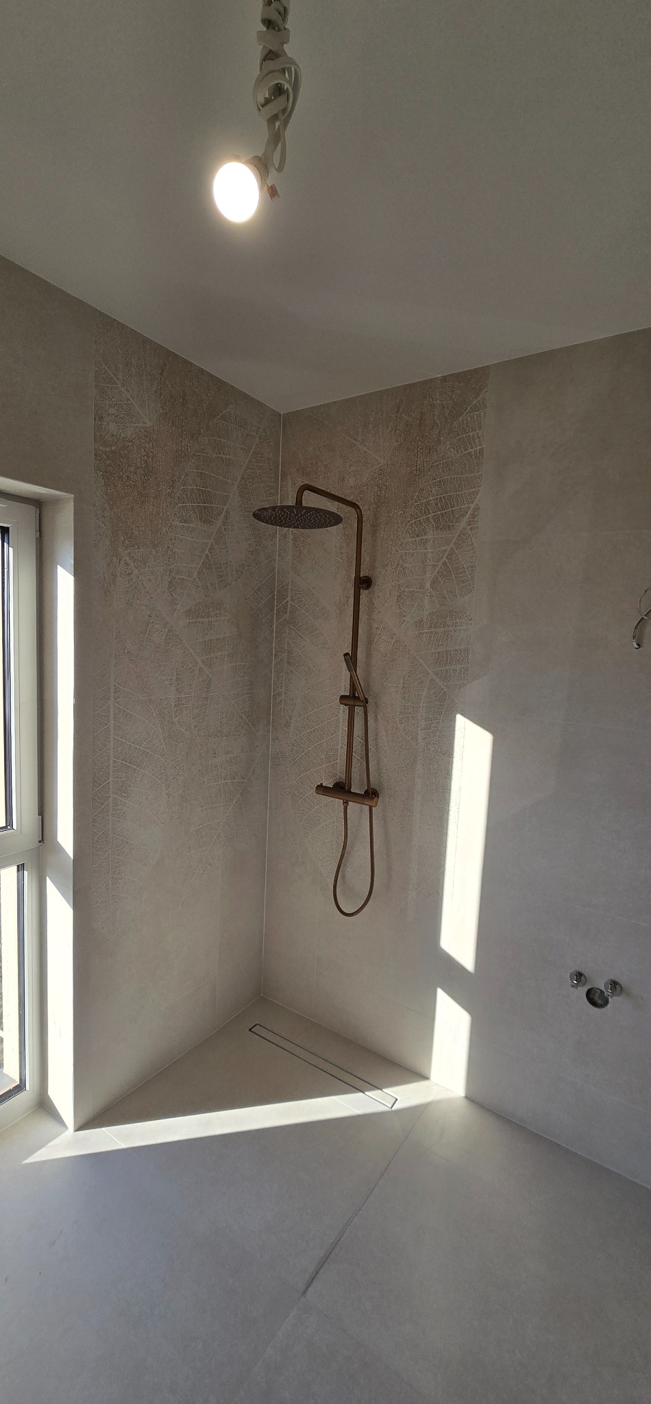 Wykończona kabina prysznicowa z beżowymi płytkami w liście, brązową armaturą i odpływem liniowym. Widoczne mocowania baterii. W rogu okno i światło słoneczne.