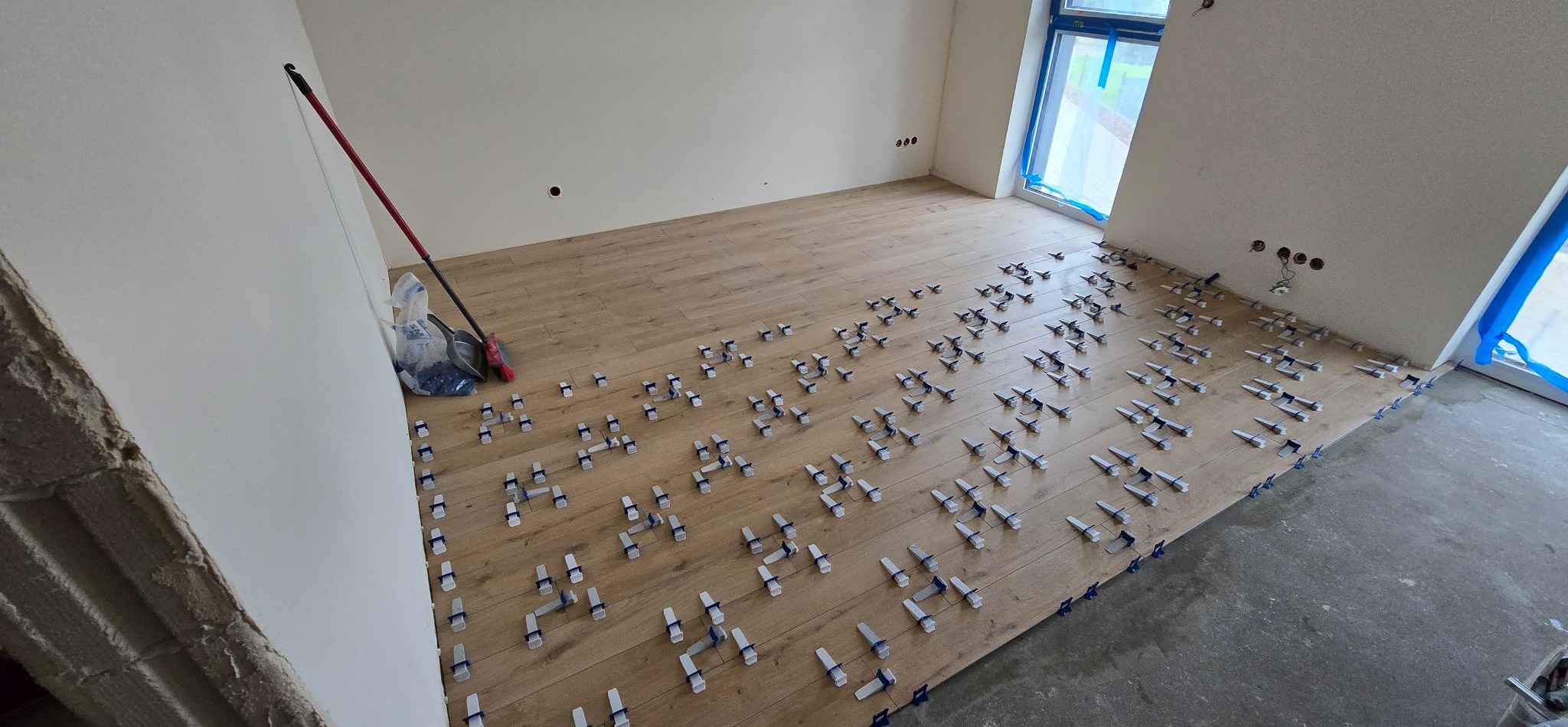 Układanie drewnopodobnych płytek podłogowych z użyciem klipsów poziomujących w jasnym pomieszczeniu. Widoczne narzędzia i worki z klipsami.