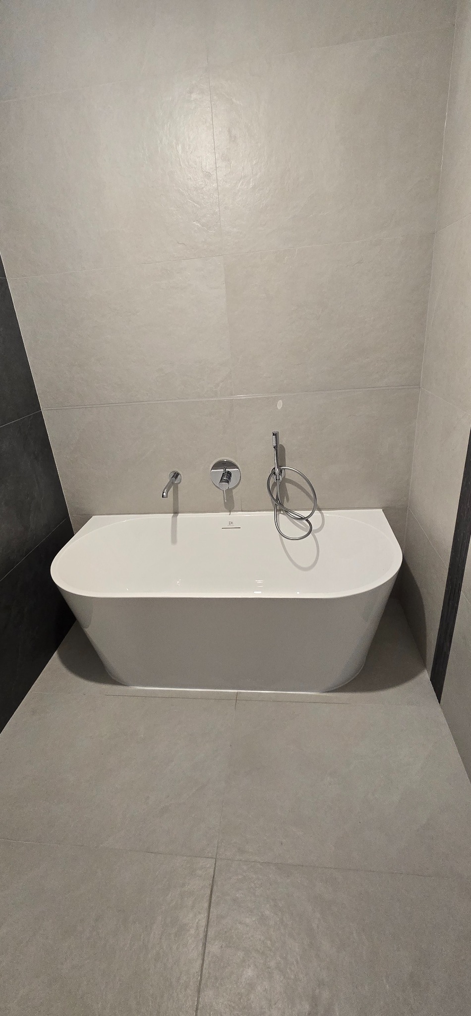 Nowoczesna, wolnostojąca wanna z chromowaną armaturą na tle szarych, matowych płytek w minimalistycznej łazience. Ujęcie centralne, podkreślające czystość i prostotę.