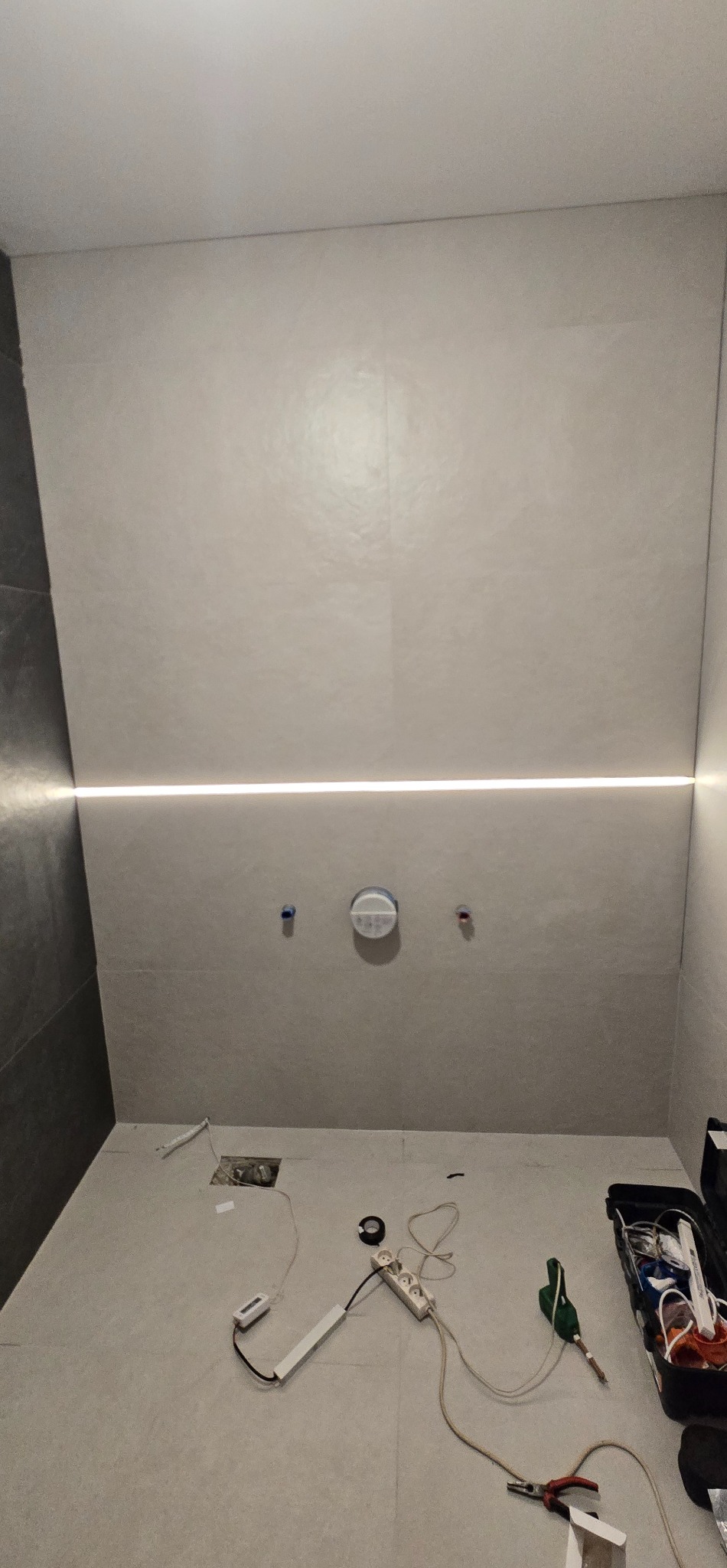 Łazienka w trakcie wykończenia: szare płytki, poziome oświetlenie LED, przygotowane instalacje, narzędzia i akcesoria na podłodze. Widok z góry.