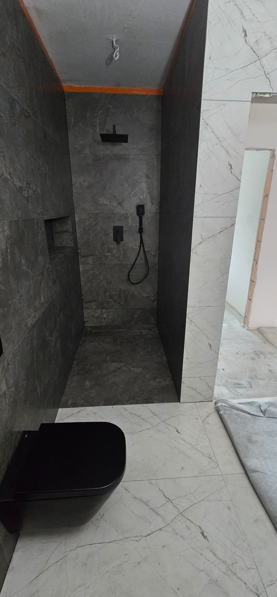 Nowoczesna kabina prysznicowa walk-in z czarną armaturą i wiszącą toaletą. Szare płytki imitujące kamień na ścianach, białe marmurowe płytki na podłodze.