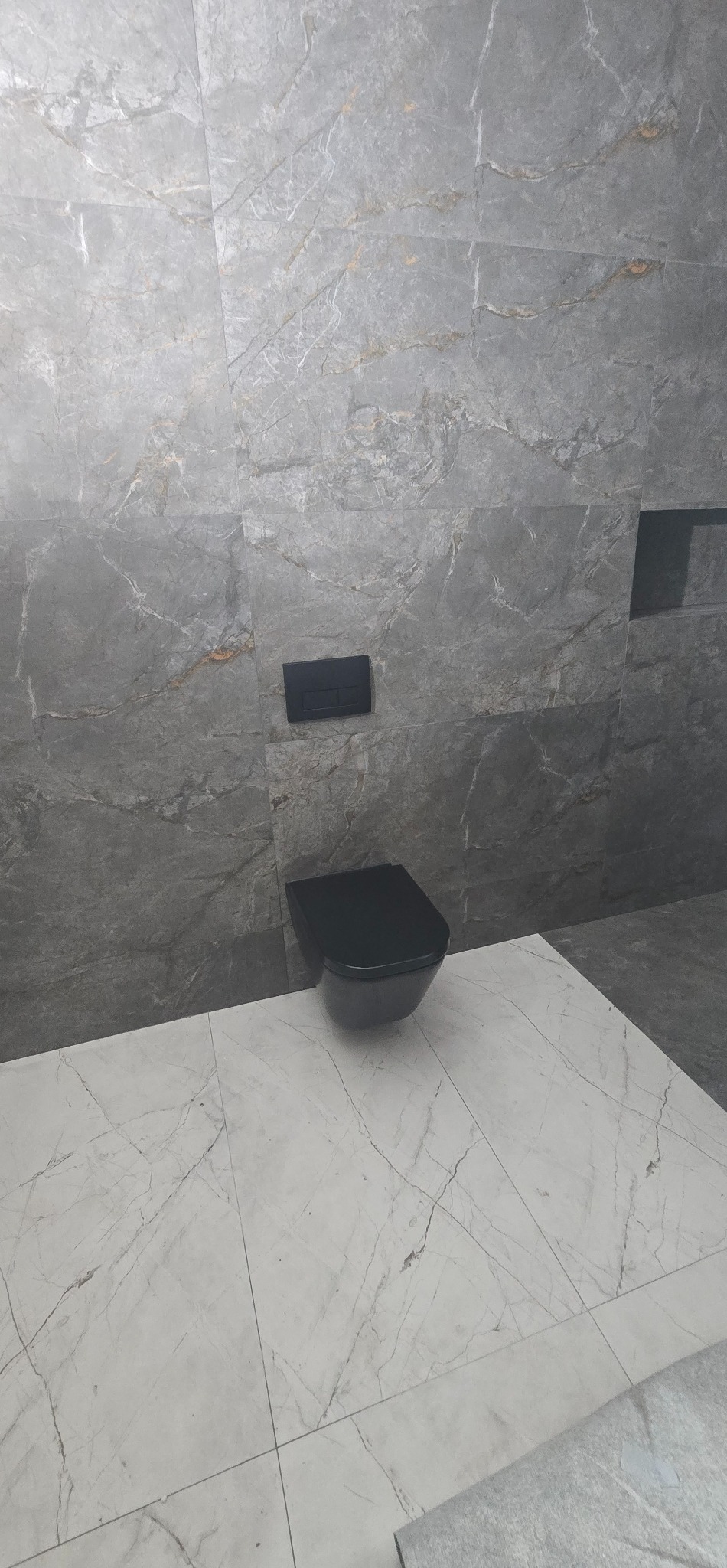 Nowoczesna łazienka z szarymi płytkami imitującymi marmur na ścianach i białymi na podłodze. Wisząca, czarna toaleta i minimalistyczny przycisk spłukujący.