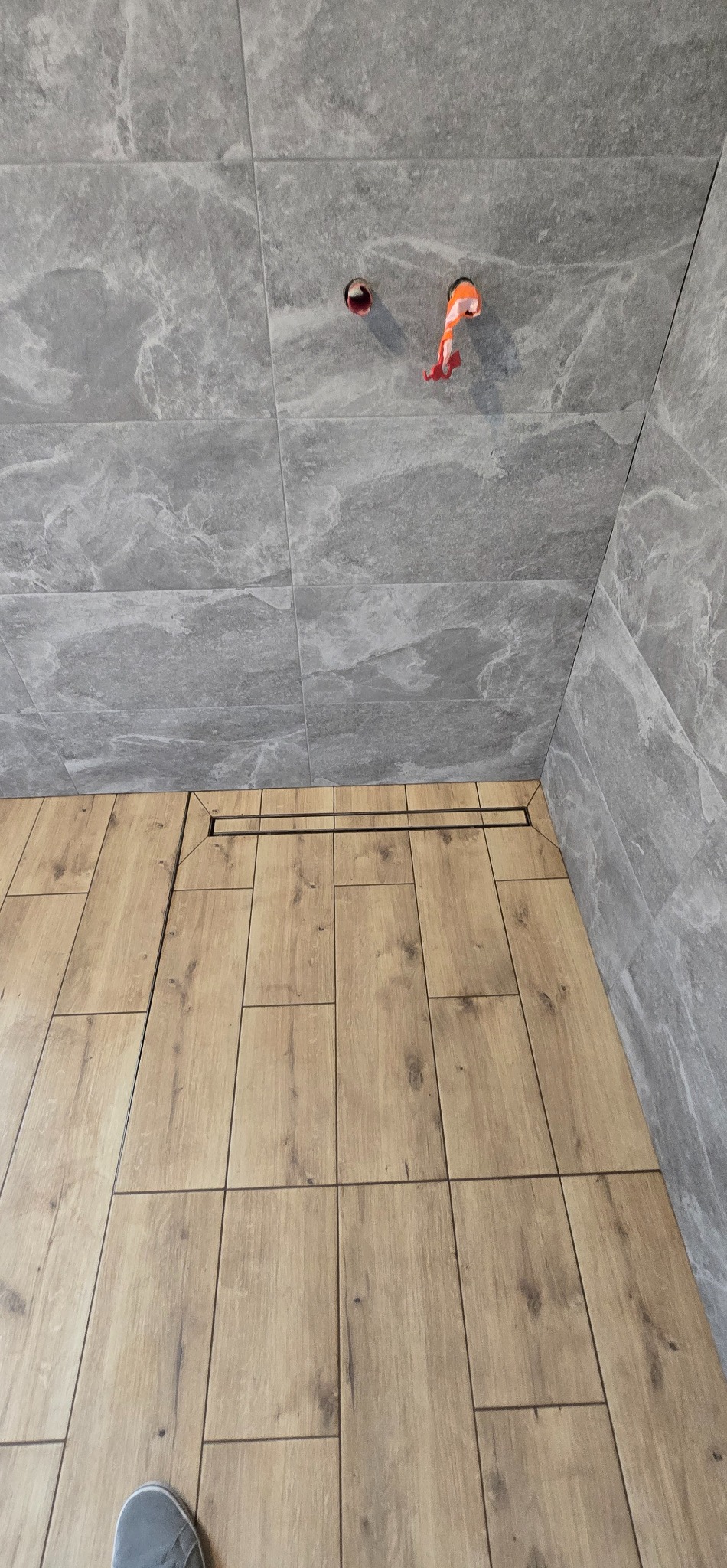 Łazienka po remoncie: drewnopodobne płytki podłogowe z odpływem liniowym, szare płytki ścienne imitujące kamień, widoczne otwory na instalacje.