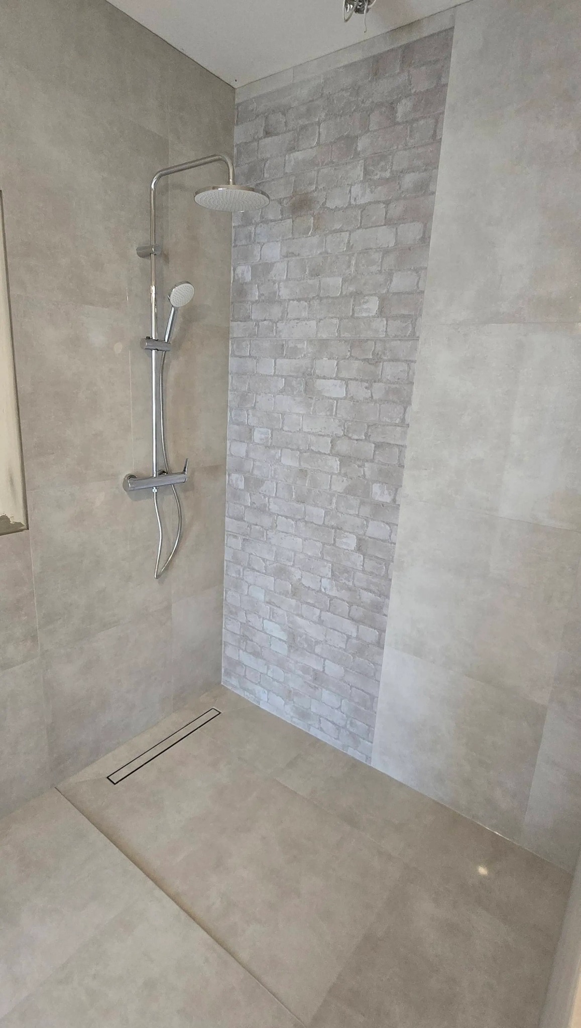 Beżowa kabina prysznicowa z odpływem liniowym i imitacją cegły na ścianie. Nowoczesny design, neutralna kolorystyka, chromowana armatura.