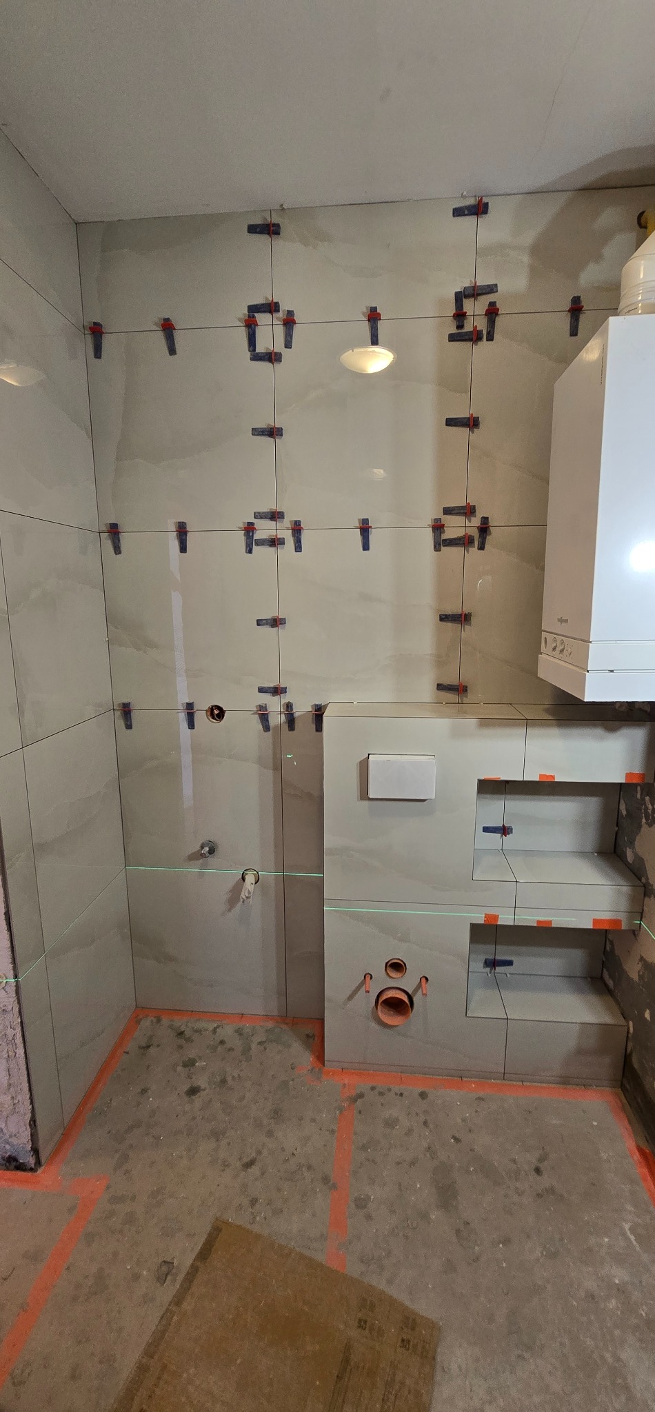 Łazienka w trakcie prac: jasne płytki na ścianach, poziomica laserowa, system poziomowania płytek. Widoczny bojler i zabudowa geberitu z półkami.