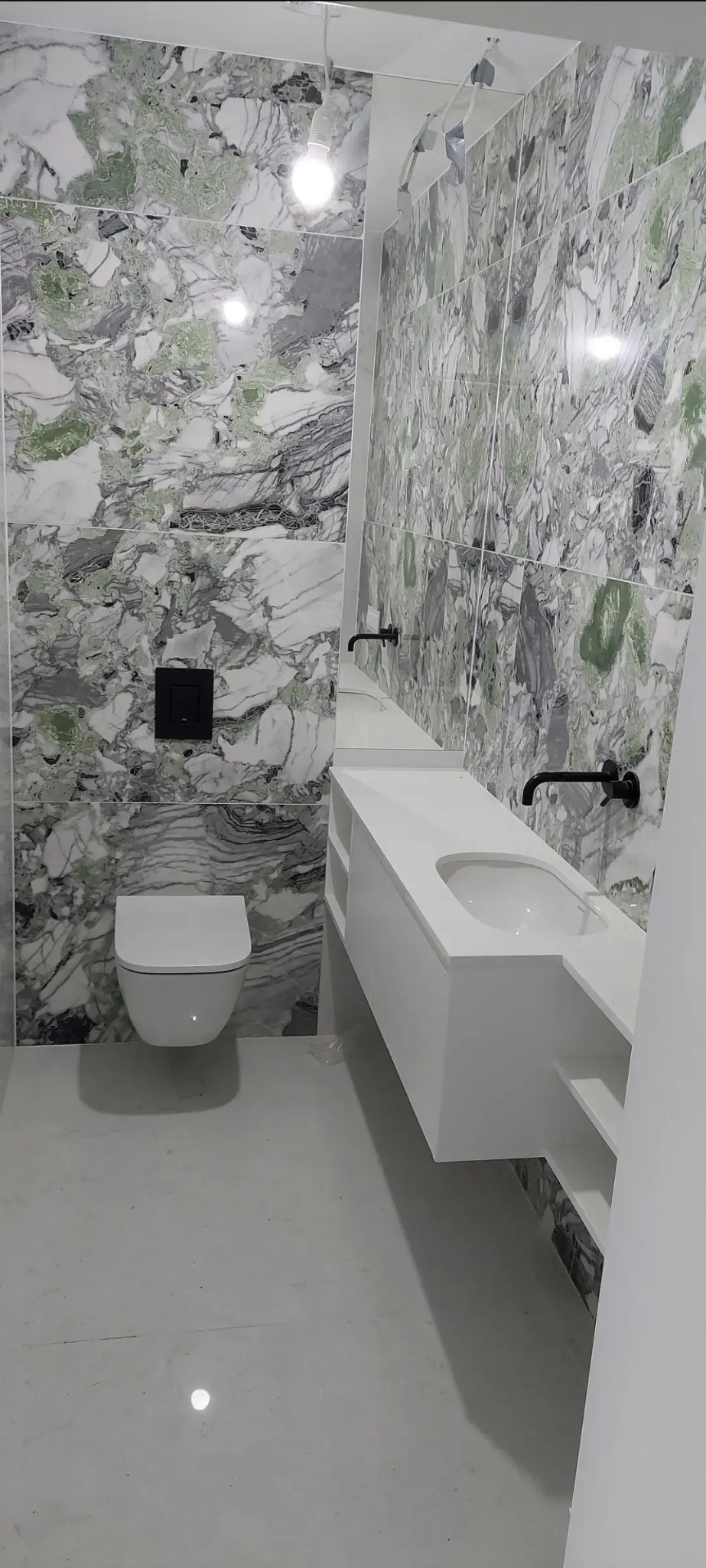 Nowoczesna łazienka z marmurowymi płytkami w odcieniach zieleni i szarości. Biała, podwieszana toaleta i umywalka z czarną armaturą. Minimalistyczny design.