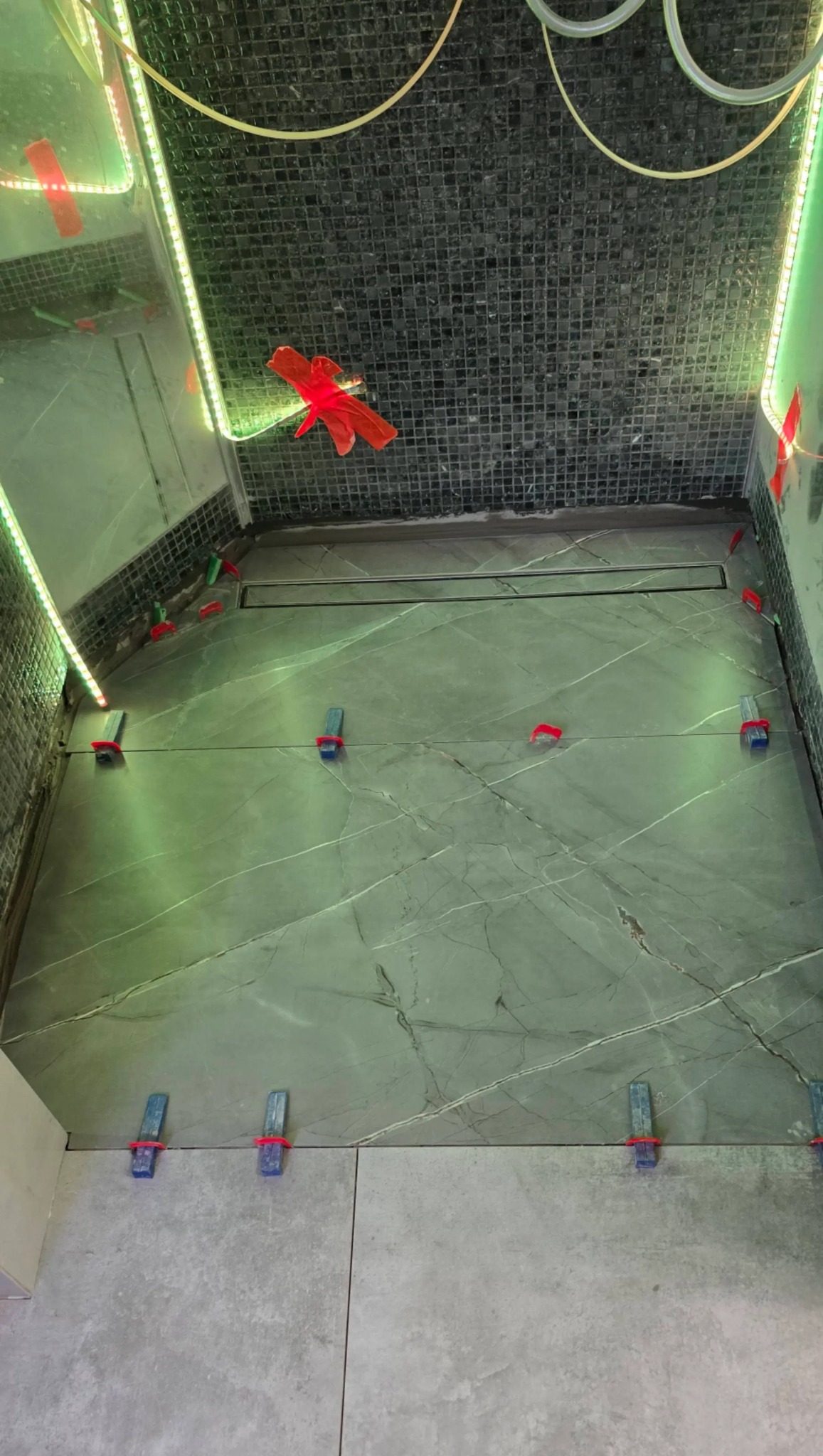 Łazienka w trakcie wykańczania: ułożone płytki podłogowe z szarym wzorem marmuru, krzyżyki glazurnicze, odpływ liniowy, mozaika na ścianie i oświetlenie LED.