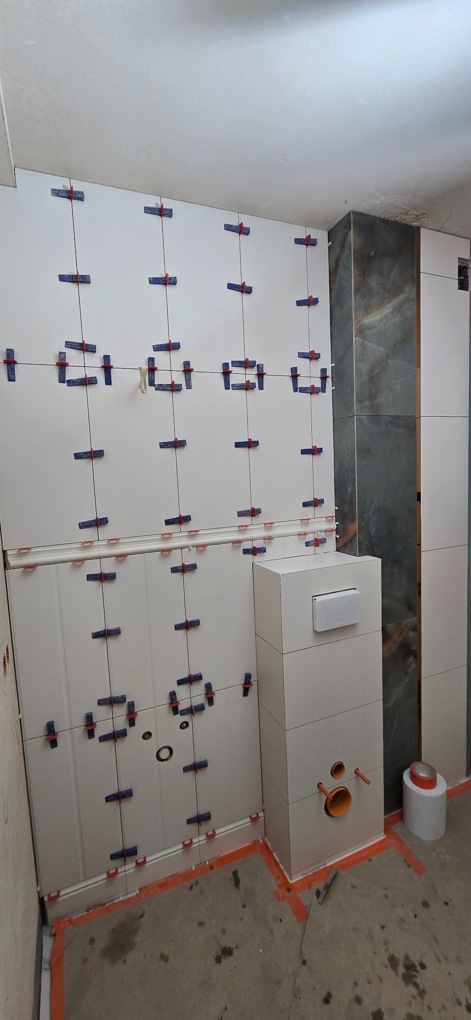 Białe płytki ścienne z niebiesko-czerwonymi krzyżykami dystansowymi w trakcie montażu. Widoczna zabudowa WC i fragment ściany z ciemnymi płytkami imitującymi kamień.