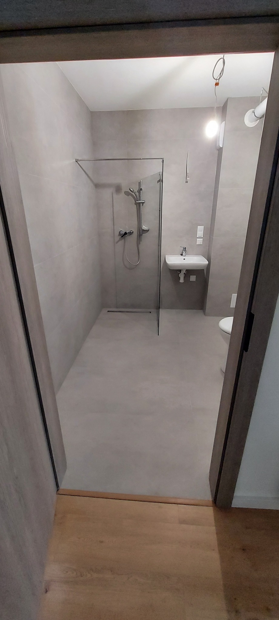 Nowoczesna łazienka w odcieniach szarości z kabiną prysznicową typu walk-in, umywalką i toaletą. Minimalistyczny design, widok zza drzwi.