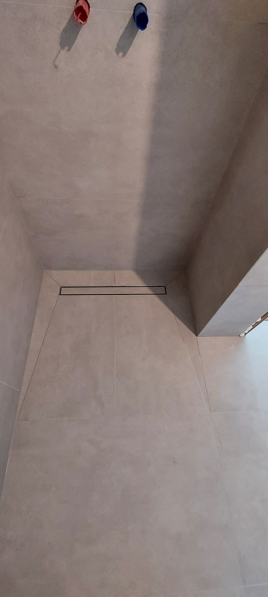 Wykończona łazienka z szarymi płytkami i odpływem liniowym. Widoczne rury doprowadzające wodę. Minimalistyczny design.