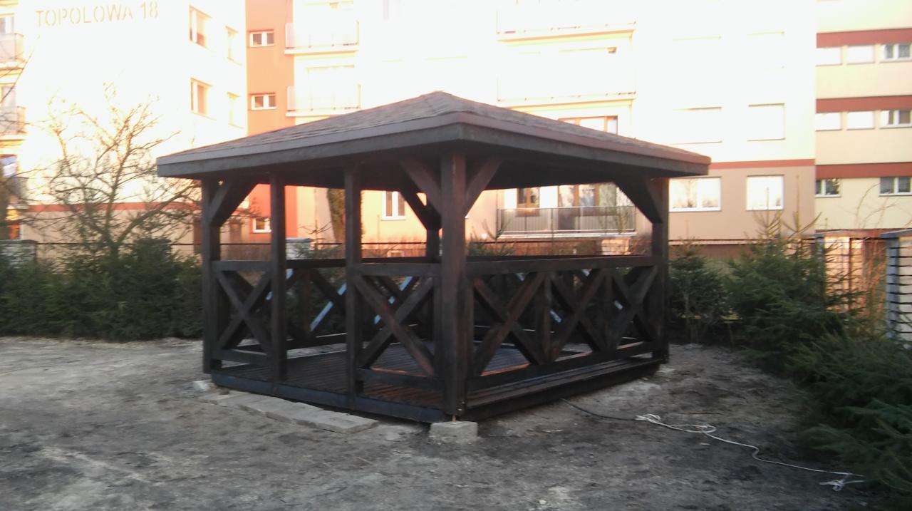 Ciemna, drewniana altana z krzyżowymi zdobieniami, ustawiona na betonowych wspornikach na ziemi, z budynkiem mieszkalnym w tle i zielenią wokół.