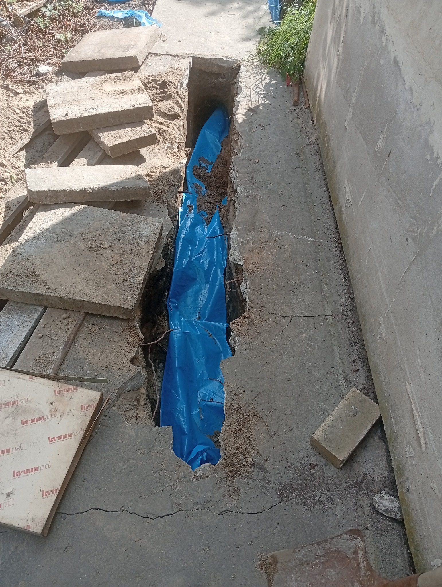 Wykop w ziemi zabezpieczony niebieską folią, obok leżą betonowe płyty chodnikowe. Widoczne ślady prac ziemnych i przygotowanie do instalacji podziemnej.