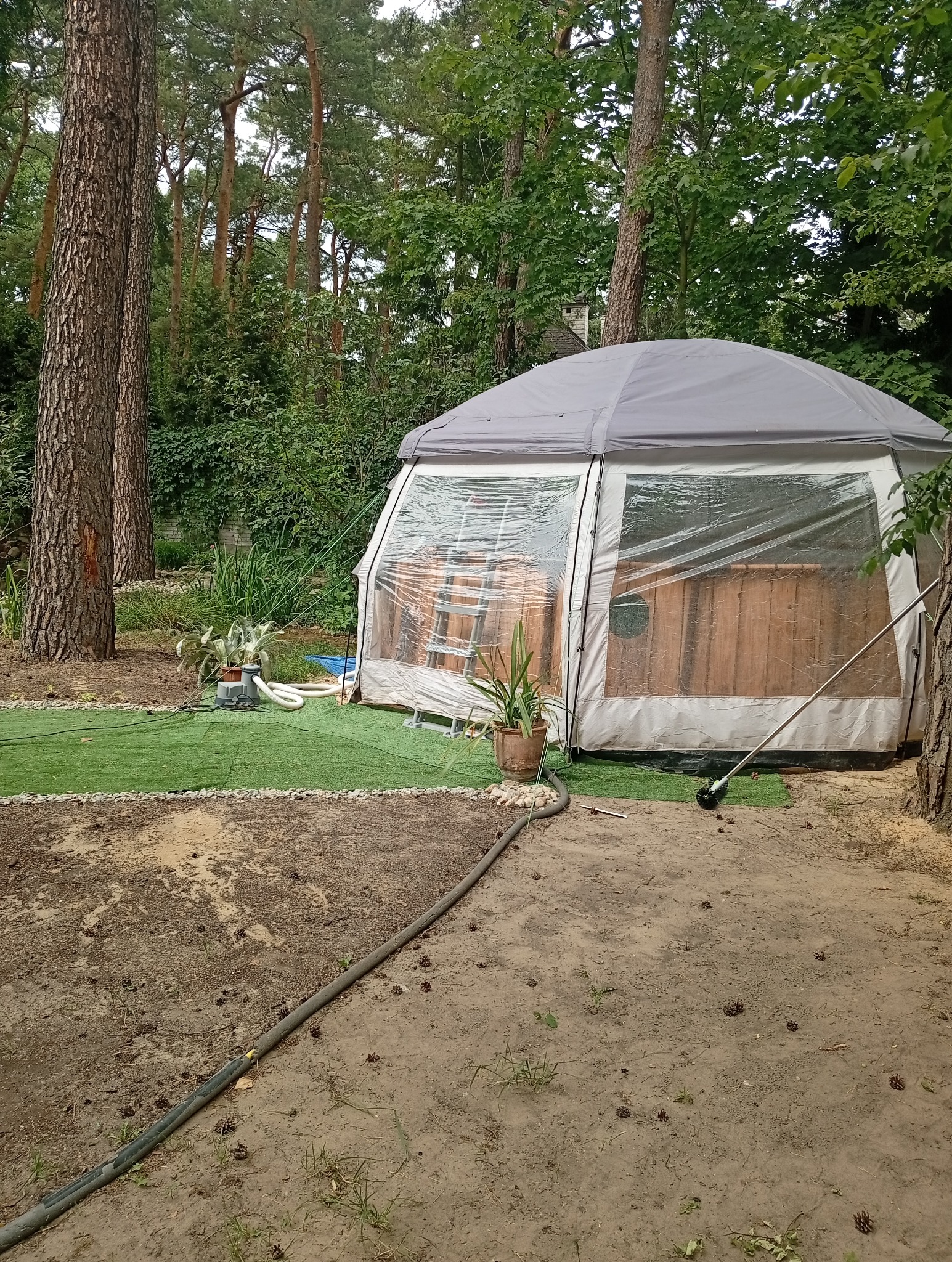 Zewnętrzna, drewniana balia w szarym namiocie z przezroczystymi ścianami, otoczona zielenią i drzewami. Wokół leżą szyszki, a obok namiotu stoi roślina w doniczce.