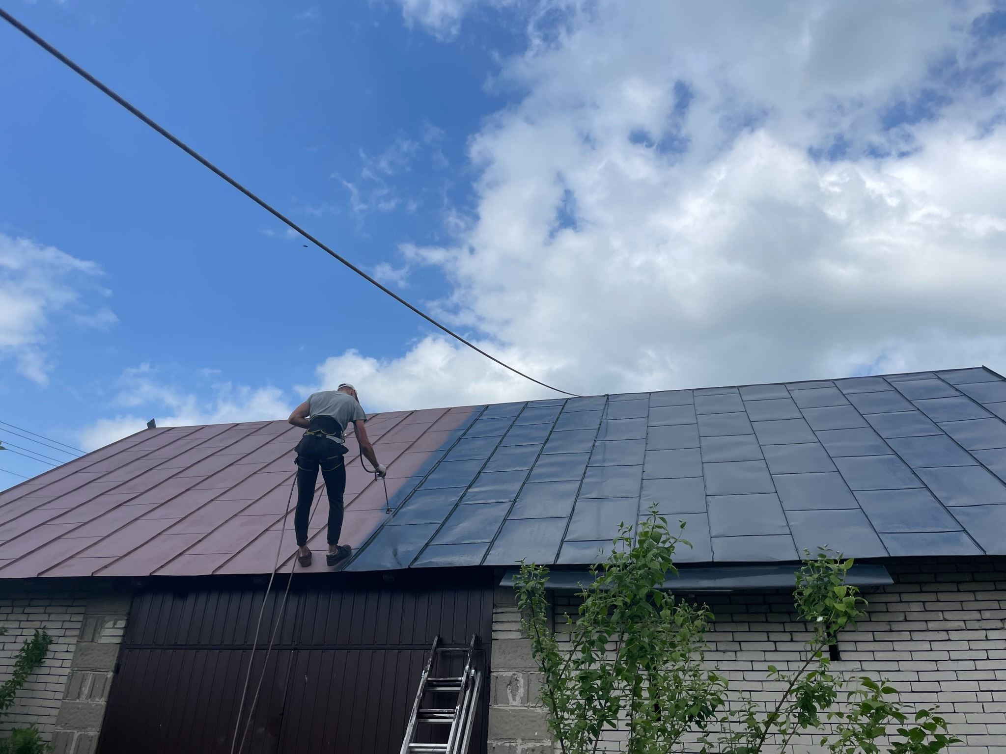 Pracownik z zabezpieczeniem linowym maluje dach z blachy trapezowej na ciemny kolor. Część dachu jest jeszcze w kolorze ceglastym. Widoczne rusztowanie i budynek.