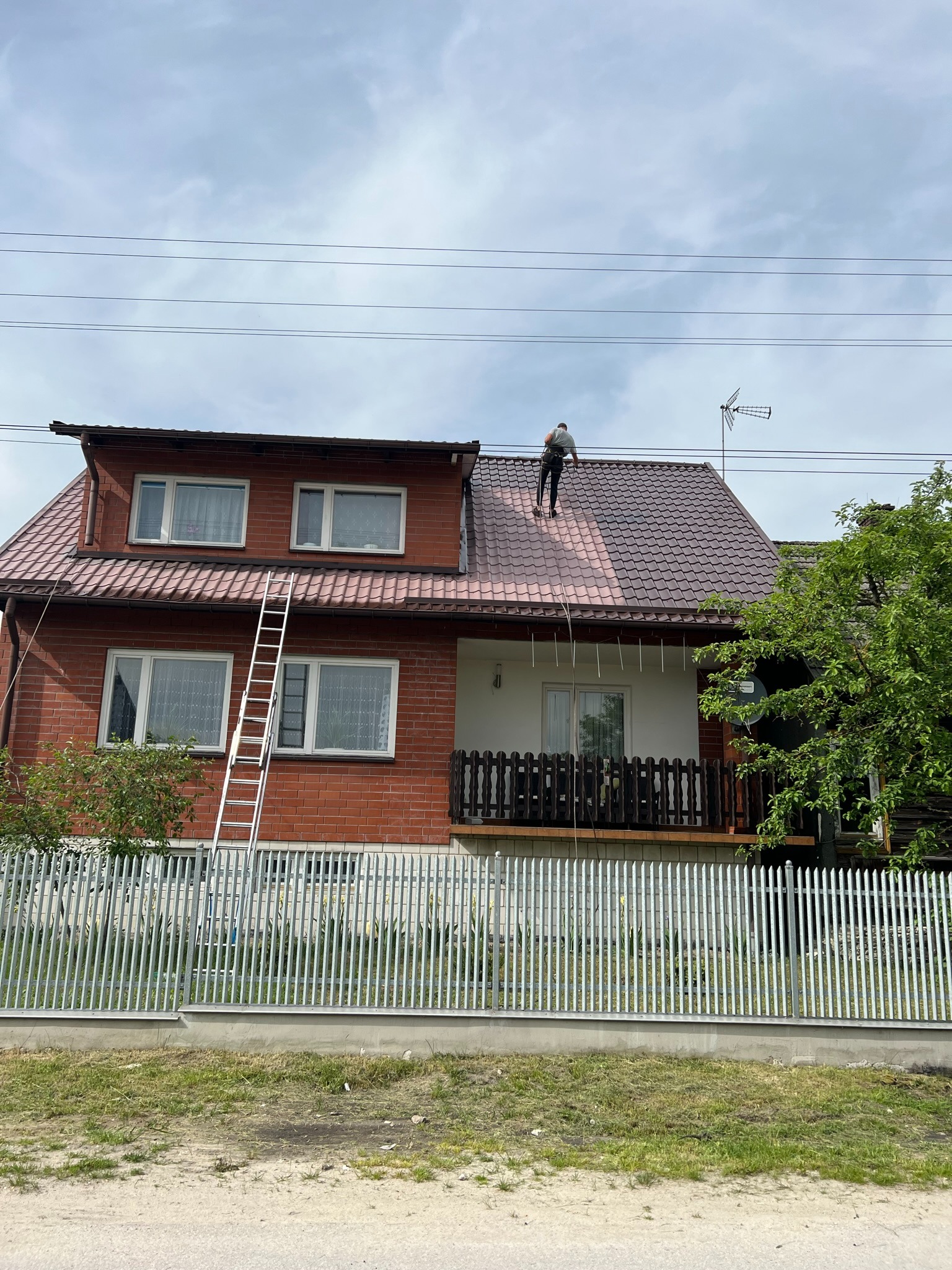 Pracownik na dachu dwukondygnacyjnego domu z cegły, częściowo pokrytym nową, brązową dachówką. Widoczna drabina oparta o ścianę i antena na dachu.