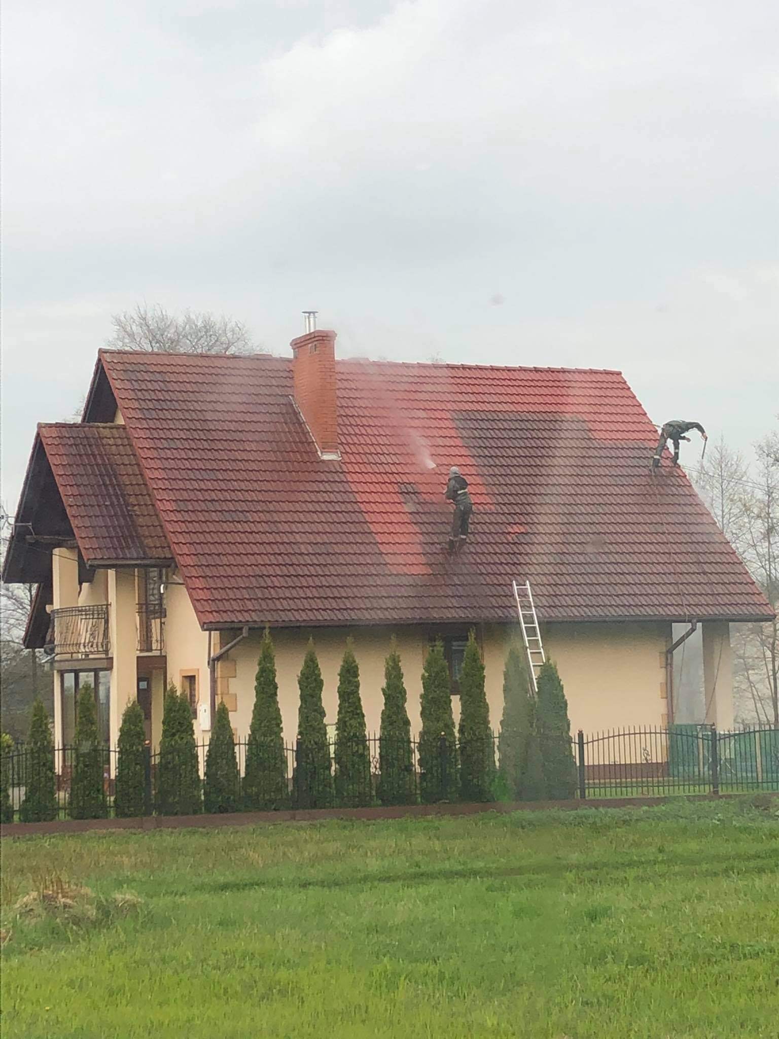 Dwóch pracowników w kombinezonach czyści dach z czerwonej dachówki za pomocą myjek ciśnieniowych. Część dachu jest już umyta, widoczna różnica w kolorze.