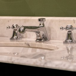 GRANITY SZCZEPAŃSKI - FILIP SZCZEPAŃSKI - Elegancka umywalka z blatem z jasnego kamienia z wyraźnym użyleniem, chromowana bateria w stylu retro, zielone tło. Detal wykończenia łazienki.