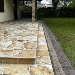 GRANITY SZCZEPAŃSKI - FILIP SZCZEPAŃSKI - Kamienny taras z nieregularnych płyt i schodami, z kolumną z kamienia. Obok trawnik i ścieżka z kostki. Ujęcie z bliska, z widocznym logo firmy.