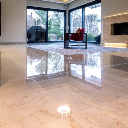 GRANITY SZCZEPAŃSKI - FILIP SZCZEPAŃSKI - Polerowana, beżowa posadzka kamienna z widocznym odbiciem okien i wyposażenia wnętrza. Nowoczesny salon z dużymi przeszkleniami i minimalistycznym oświetleniem.
