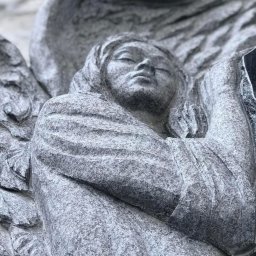 GRANITY SZCZEPAŃSKI - FILIP SZCZEPAŃSKI - Granitowa rzeźba anioła z zamkniętymi oczami, detal nagrobka. Szary kamień z widoczną fakturą. Ujęcie z dołu podkreśla monumentalność.