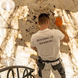 GRANITY SZCZEPAŃSKI - FILIP SZCZEPAŃSKI - Pracownik w trakcie renowacji podświetlanej ściany z marmuru o unikalnym wzorze, widoczny od tyłu, z narzędziem w dłoni, oświetlenie dekoracyjne i fragment schodów.