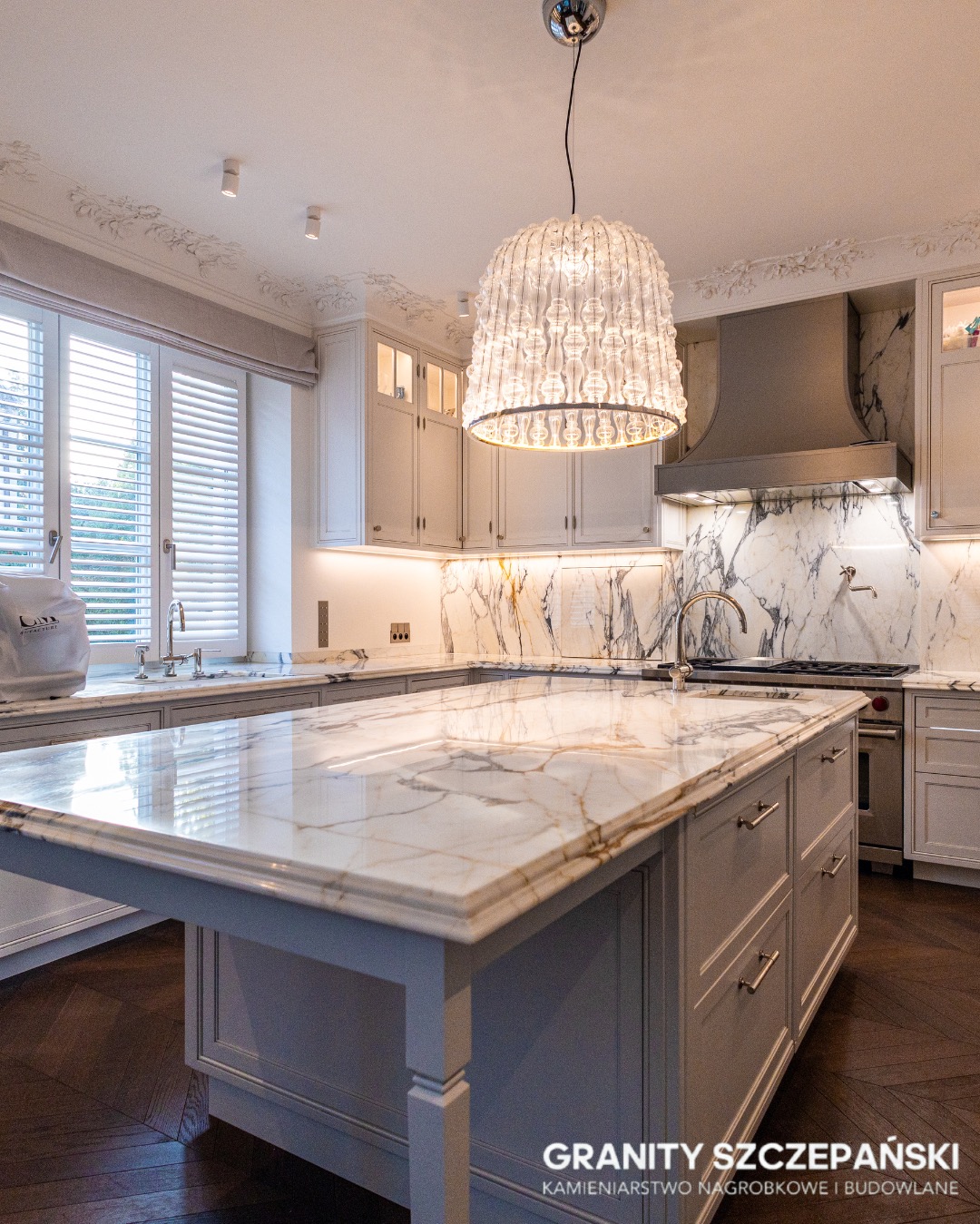 Elegancka kuchnia z wyspą i blatem z białego marmuru z złotymi żyłami, oświetlona dekoracyjną lampą. Marmur również na ścianie nad blatem. Parkiet w jodełkę.