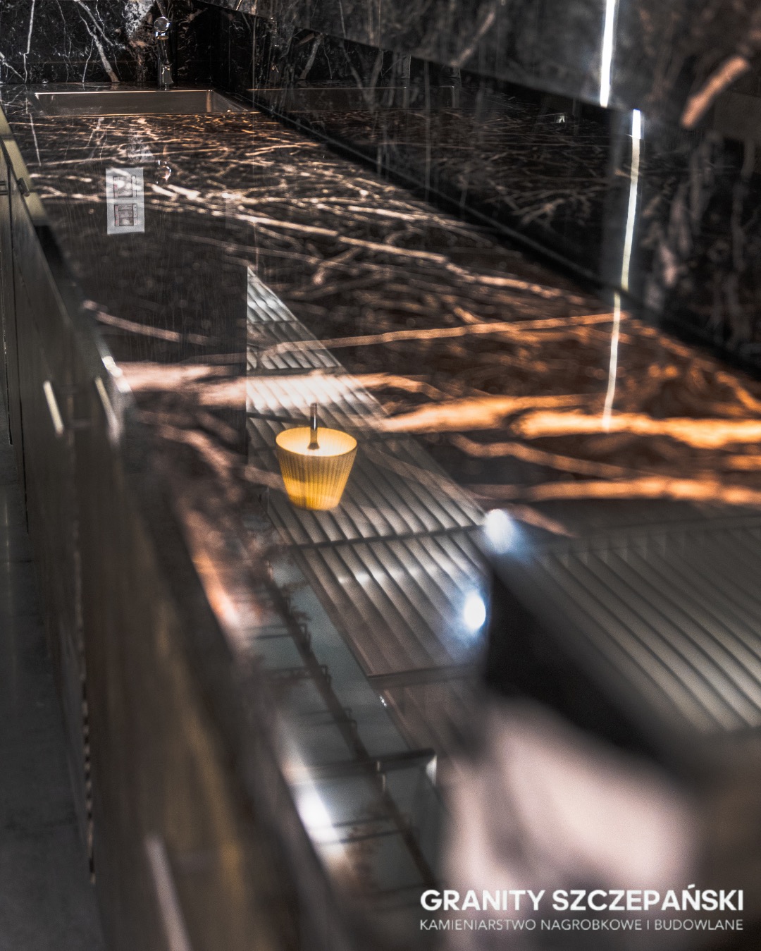 Błyszczący, czarny blat z marmurkowym wzorem, oświetlony ciepłym światłem świecy, tworzy elegancką i luksusową przestrzeń. Widoczne odbicia podkreślają gładką powierzchnię.