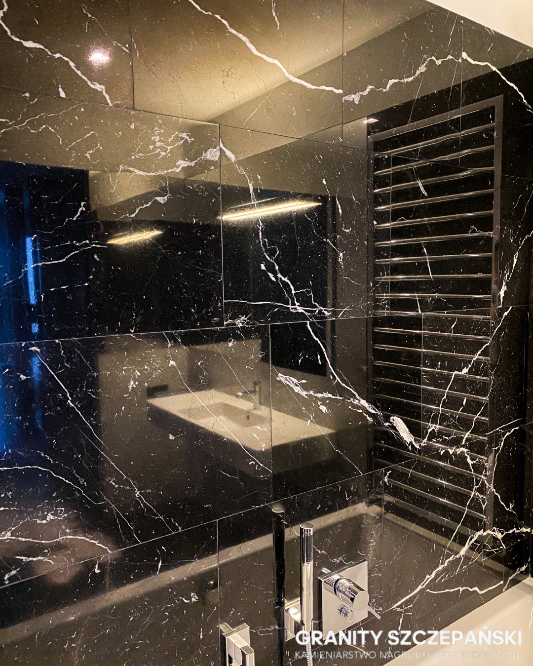 Elegancka łazienka z czarnymi płytami kamiennymi o białym żyłkowaniu, chromowana armatura i grzejnik drabinkowy. Widoczne odbicie umywalki w polerowanej powierzchni.