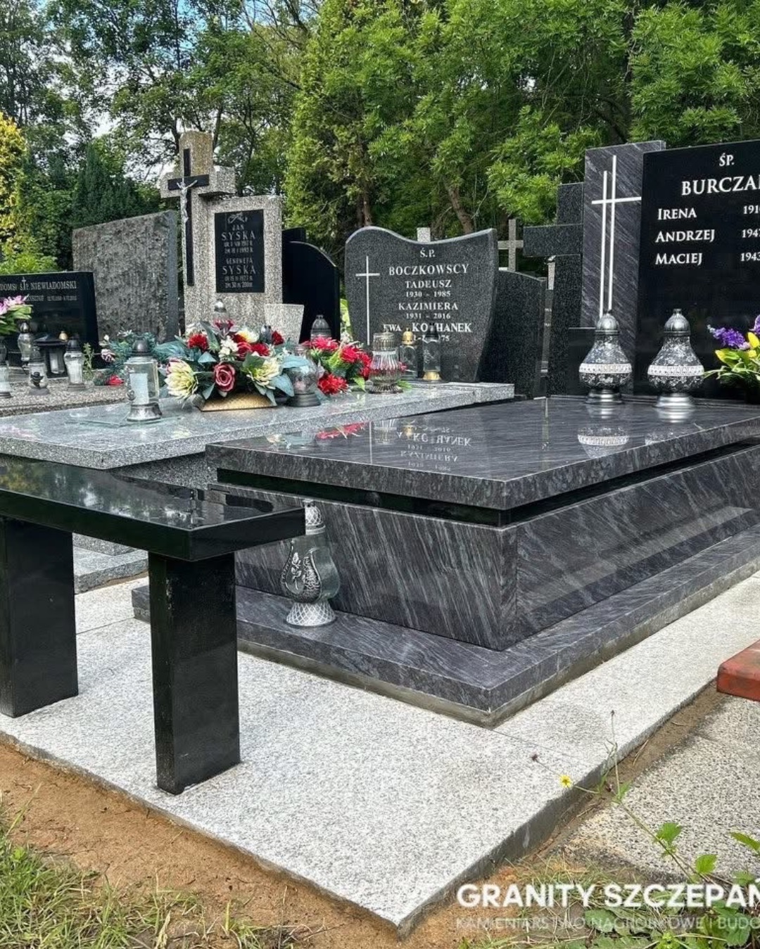 Granitowy nagrobek w odcieniach szarości z czarnym stolikiem na cmentarzu, otoczony zielenią drzew. Widoczne inskrypcje i znicze.