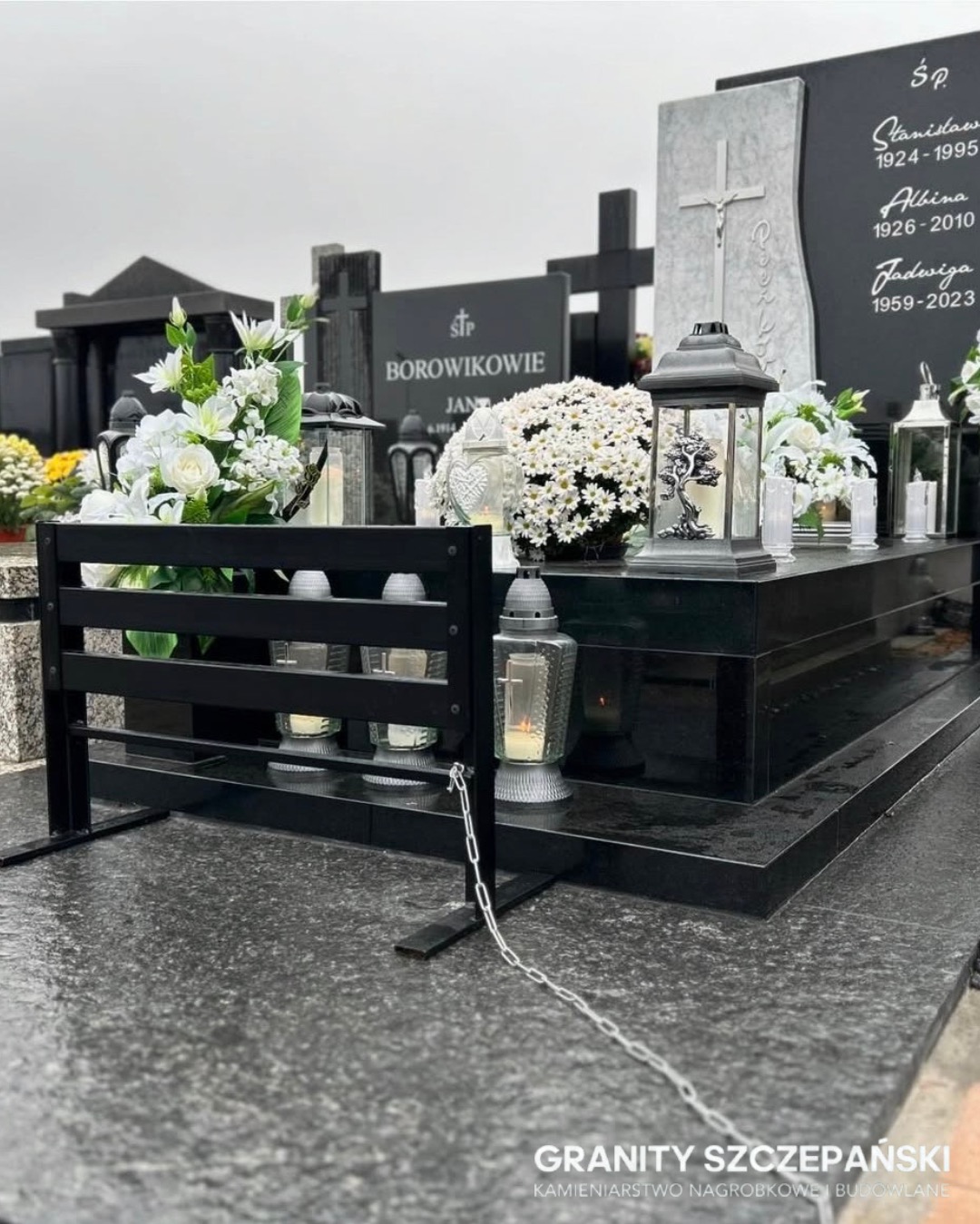 Czarny, granitowy nagrobek z kwiatami i lampionami, z widoczną metalową barierką i łańcuchem. Nagrobek z inskrypcjami i krzyżem, w pochmurny dzień.