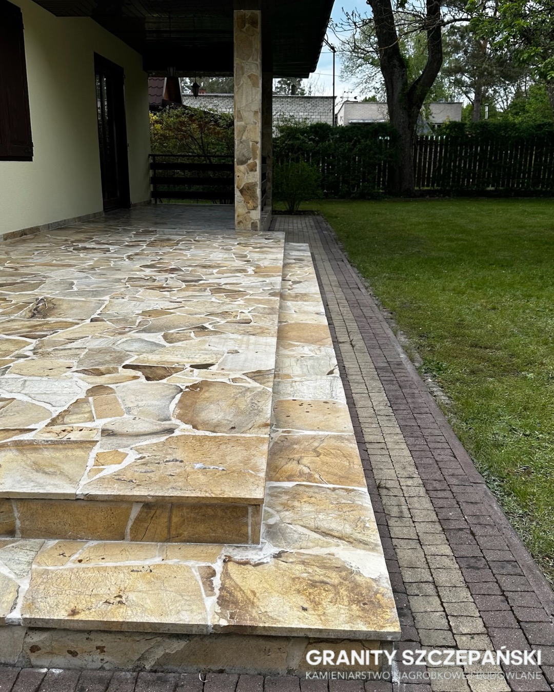 Kamienny taras z nieregularnych płyt i schodami, z kolumną z kamienia. Obok trawnik i ścieżka z kostki. Ujęcie z bliska, z widocznym logo firmy.