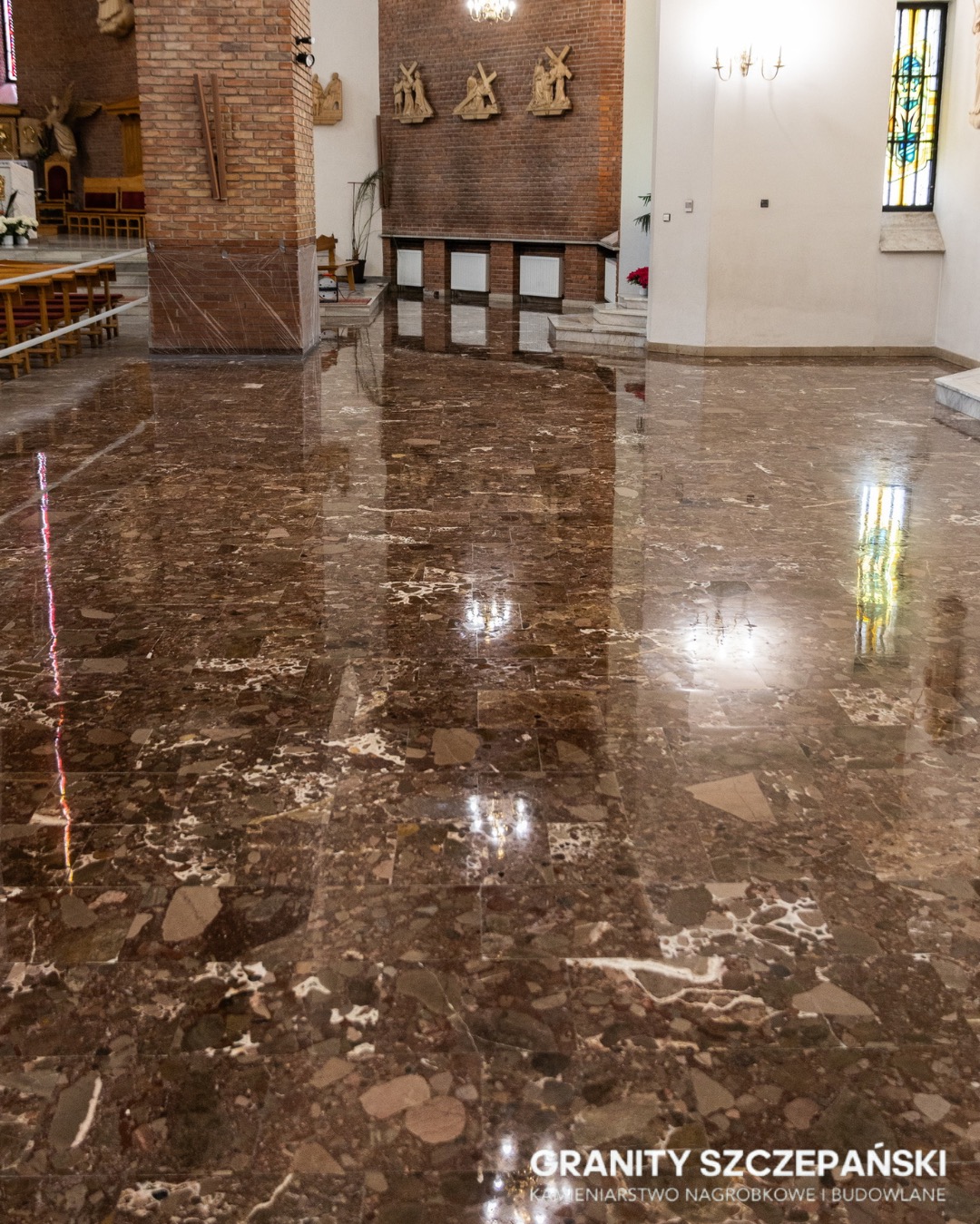 Polerowana, brązowa posadzka z kamienia naturalnego w kościele, odbijająca światło. Widoczny fragment kolumny z cegły i elementy wyposażenia wnętrza.