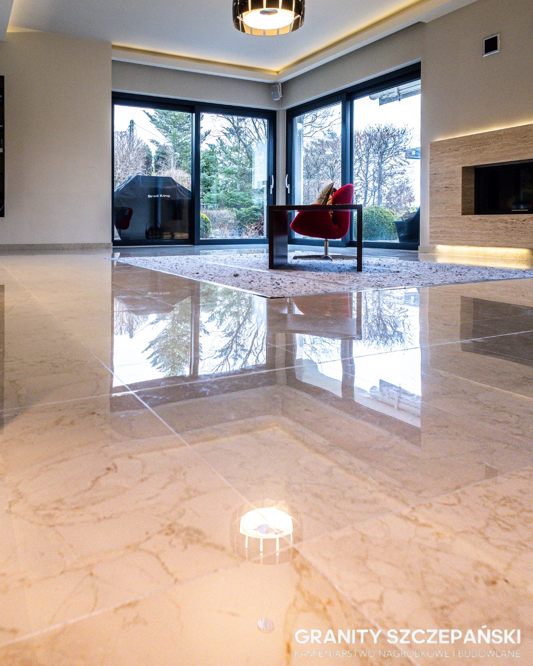 Polerowana, beżowa posadzka kamienna z widocznym odbiciem okien i wyposażenia wnętrza. Nowoczesny salon z dużymi przeszkleniami i minimalistycznym oświetleniem.